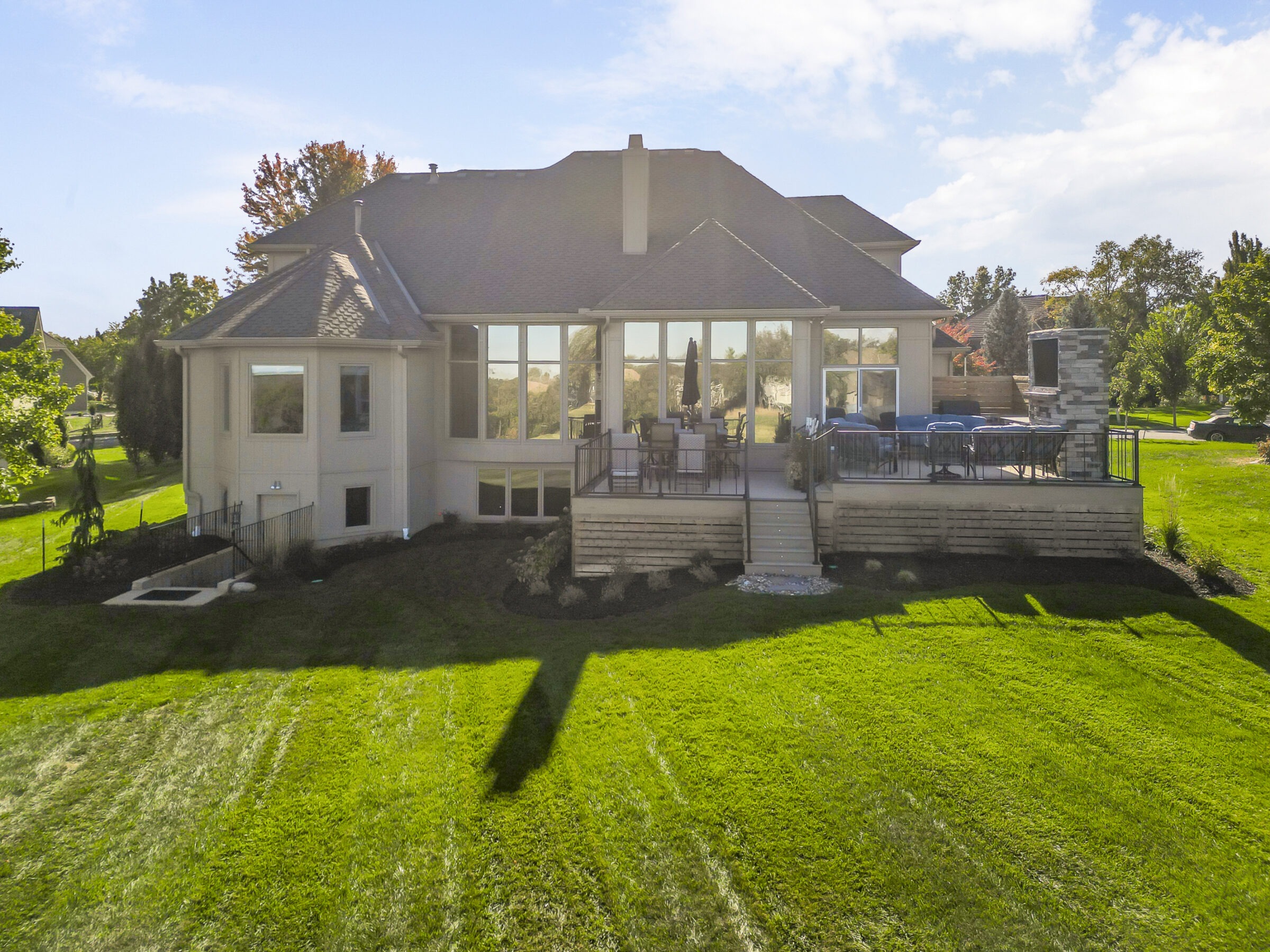 Large modern house with extensive windows, elevated deck featuring outdoor furniture, surrounded by a well-maintained lawn and trees under a clear sky.