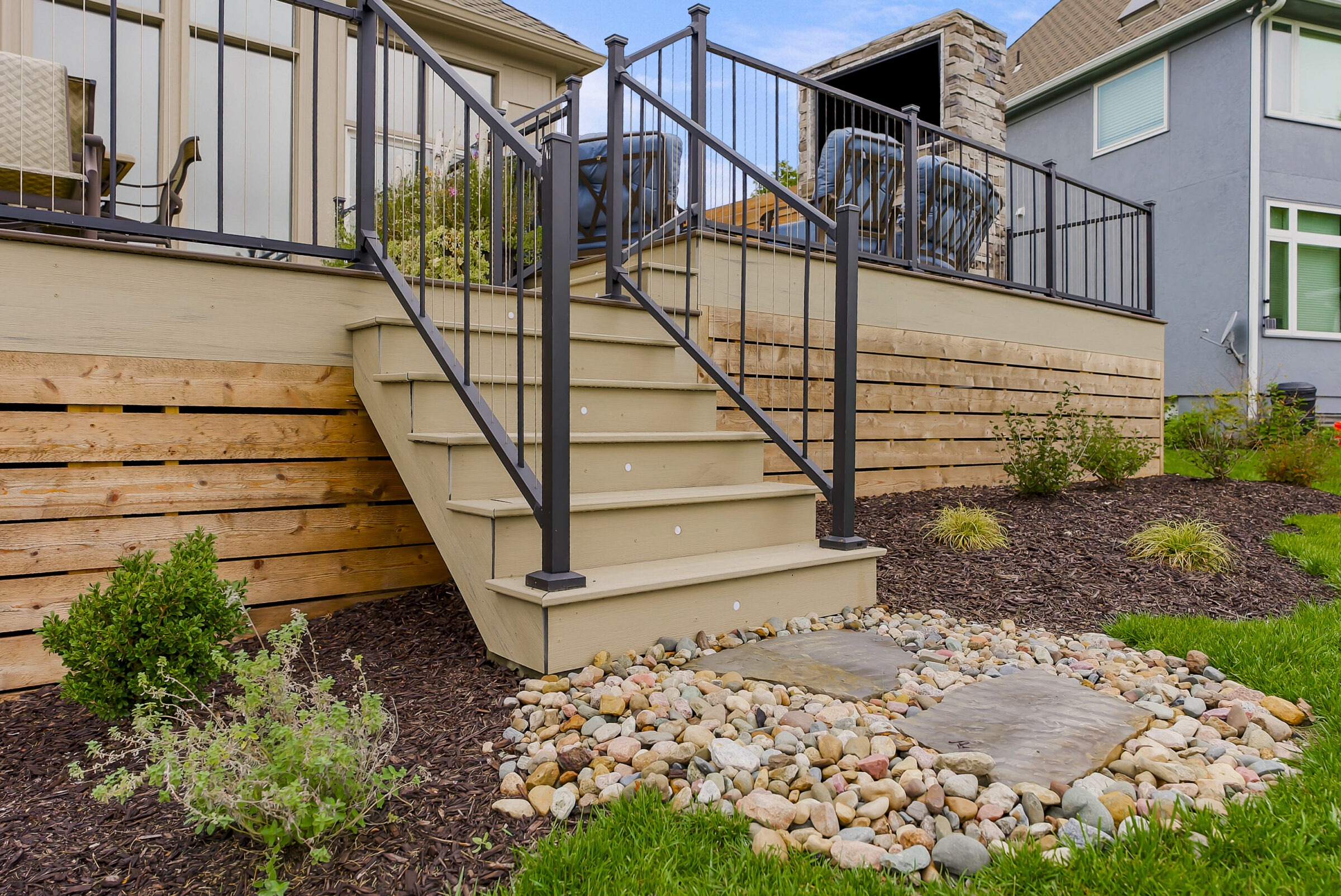 Wooden deck with black metal railing, leading to a landscaped garden with mulch, plants, and stone pathway, beside residential houses.