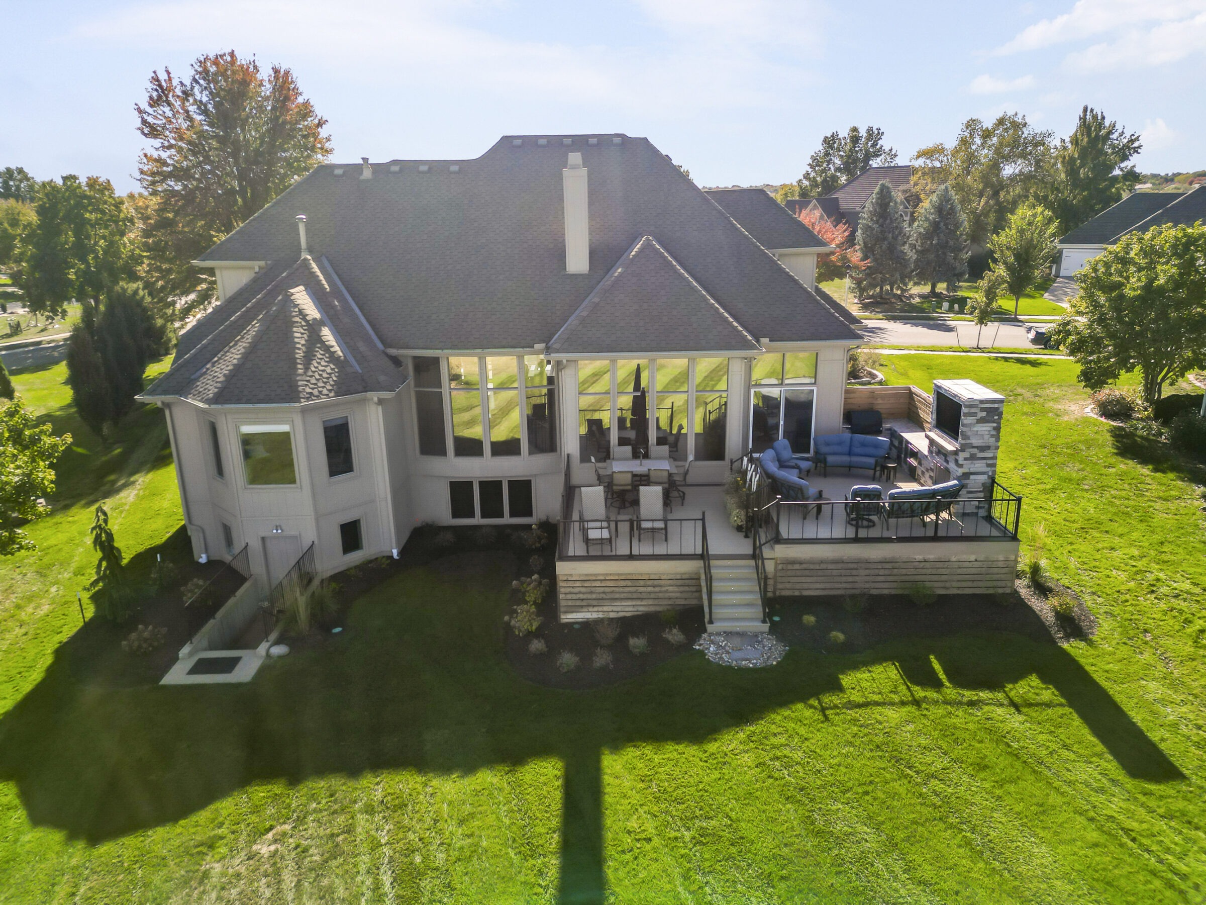 A large suburban house with a spacious deck and outdoor seating, surrounded by green lawn and trees on a sunny day.