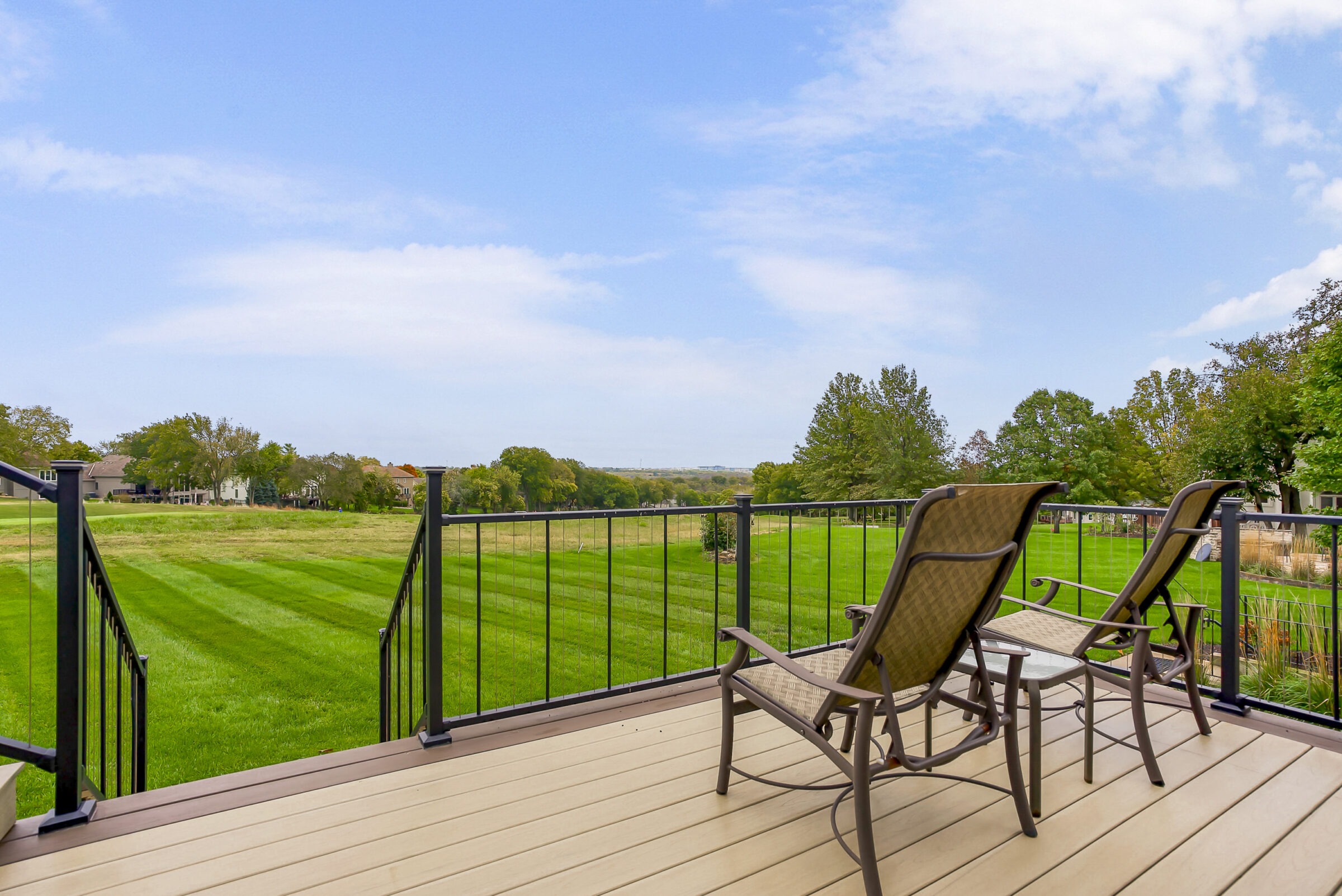 Two empty chairs on a wooden deck overlook a lush, sprawling green field under a bright blue sky with scattered clouds.