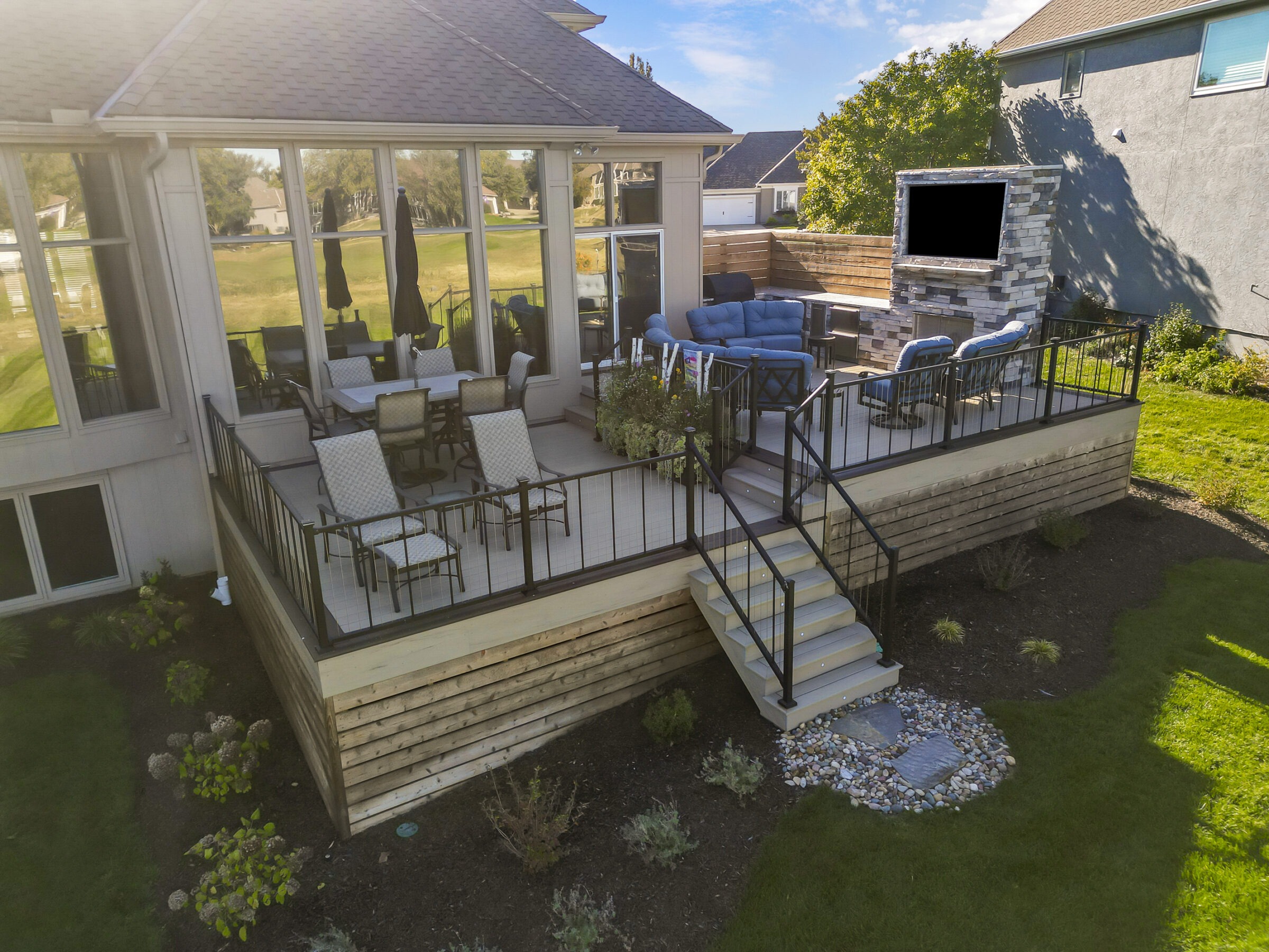 A modern backyard patio features seating and dining areas with a fireplace, surrounded by greenery, under a sunny sky.