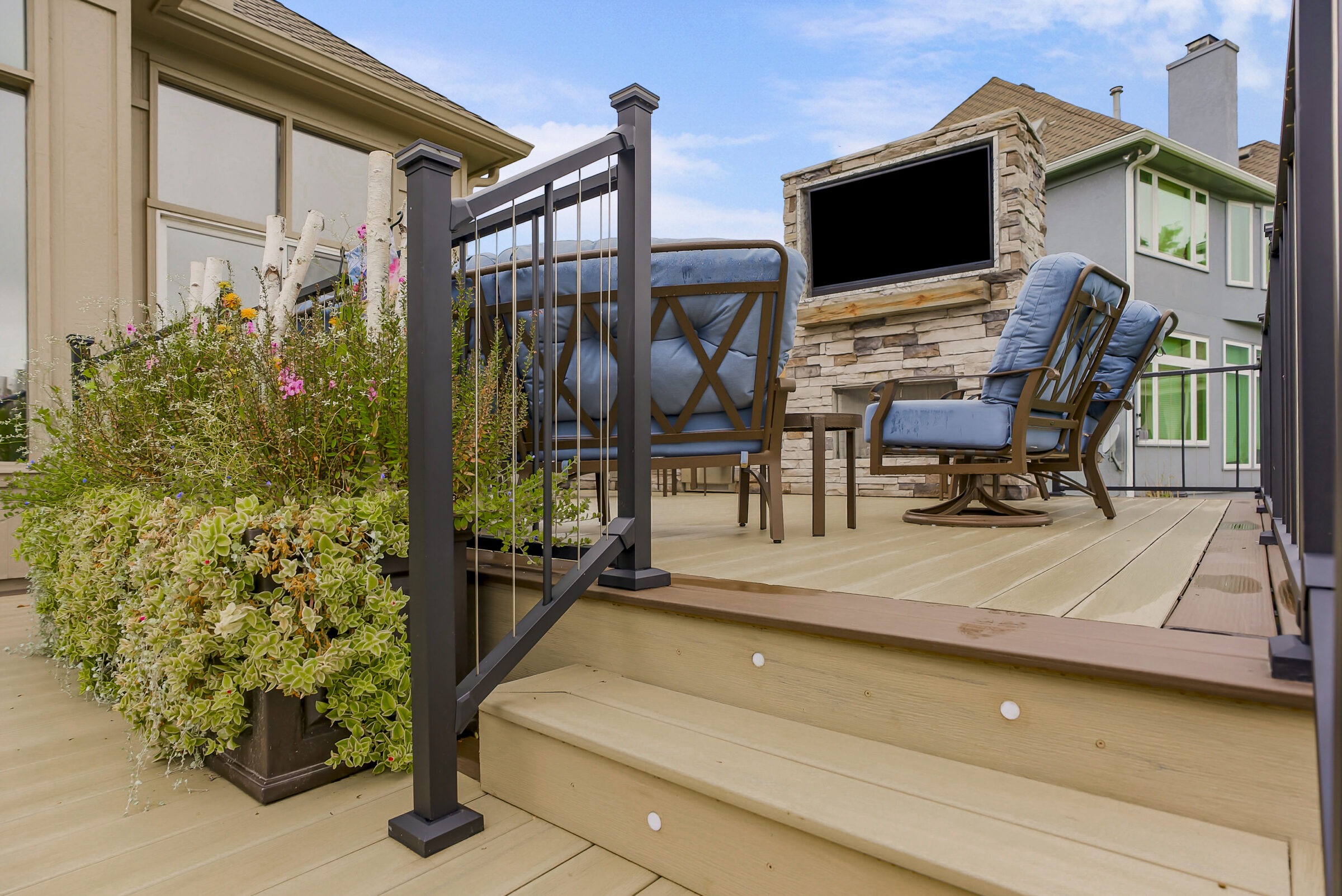 Modern patio with blue cushioned chairs, outdoor fireplace, and lush green plants. Elegant railing surrounds the wooden deck under a clear blue sky.