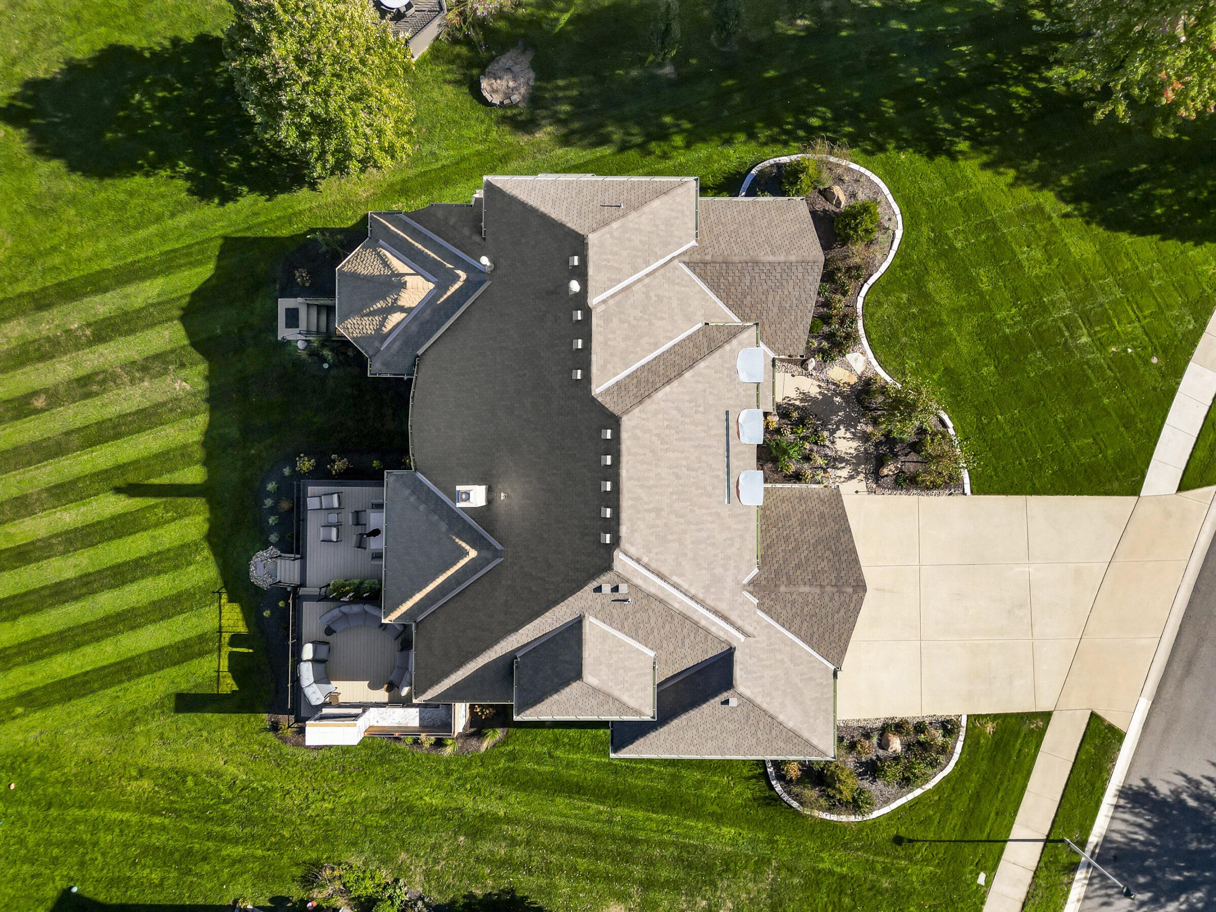 Aerial view of a residential house with landscaped gardens, driveway, and surrounding greenery on a sunny day.