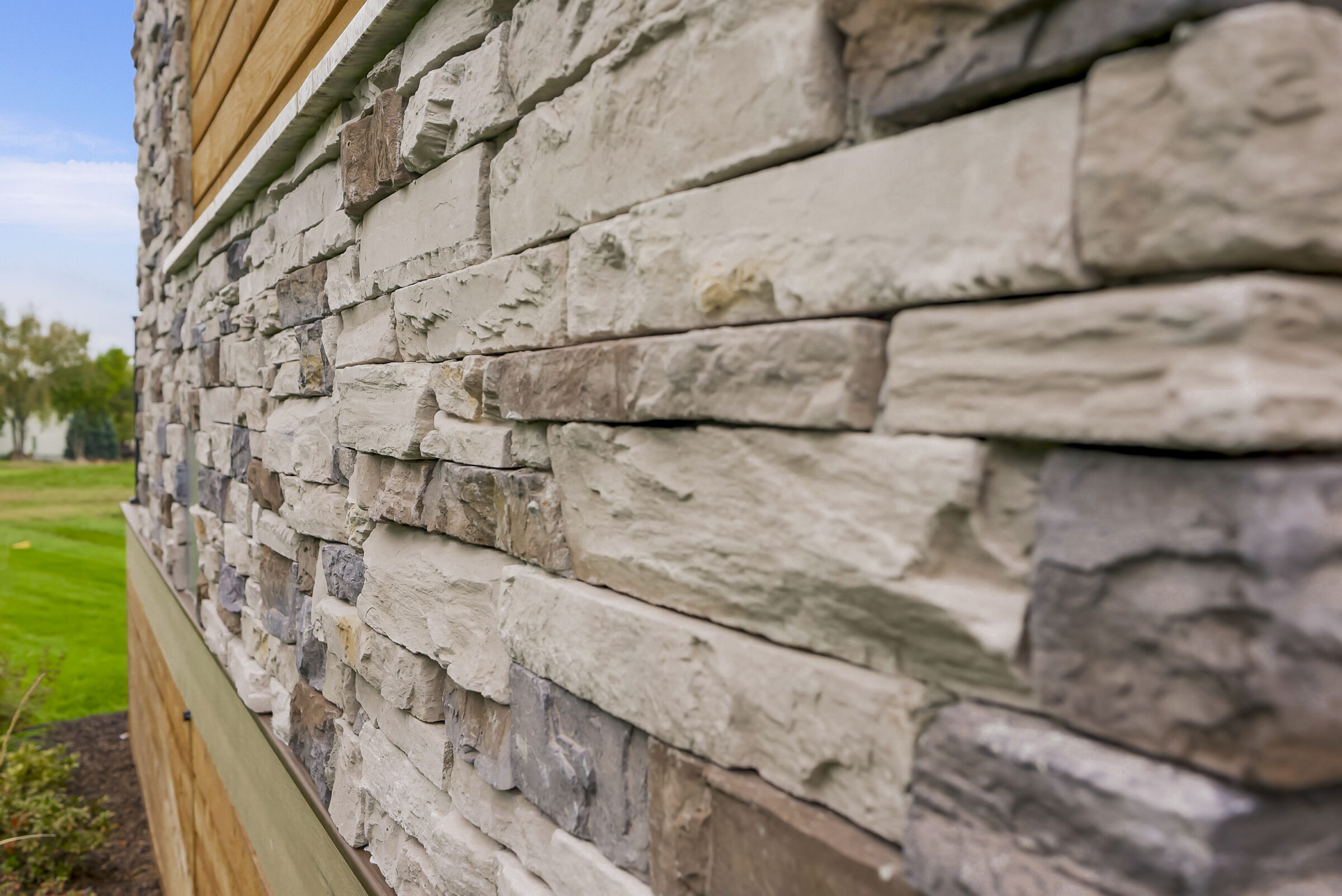 Close-up of a stacked stone wall exterior with a wooden section near a green lawn and foliage in the background.