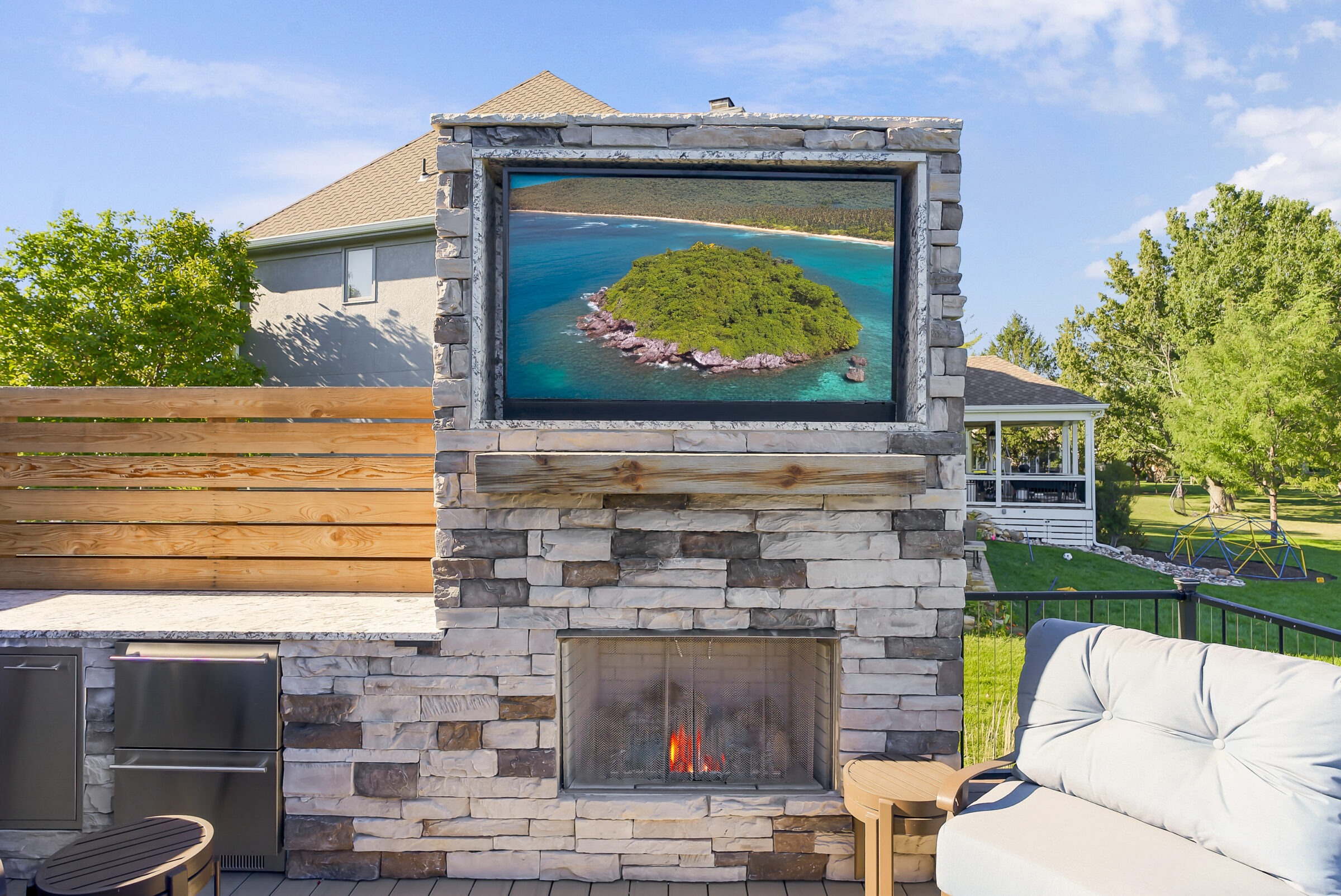 A cozy outdoor patio features a stone fireplace, comfortable seating, and a TV displaying a serene island view beneath a clear blue sky.
