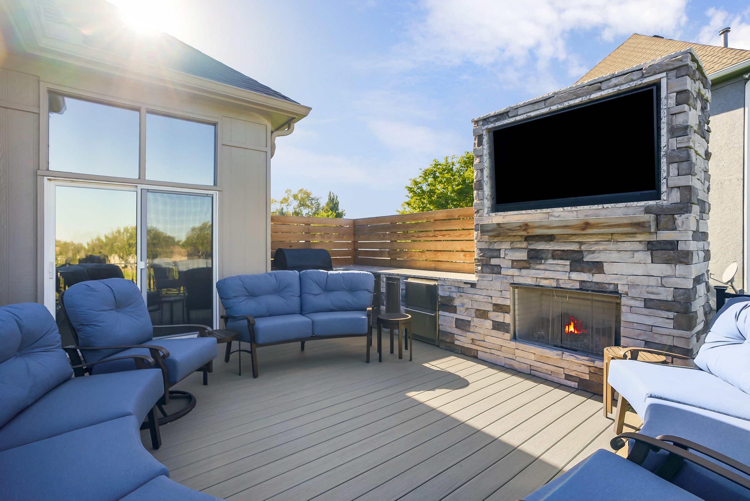 A sunny patio features blue cushioned chairs, stone fireplace, large TV, and wooden privacy fence, creating a cozy outdoor relaxation space.