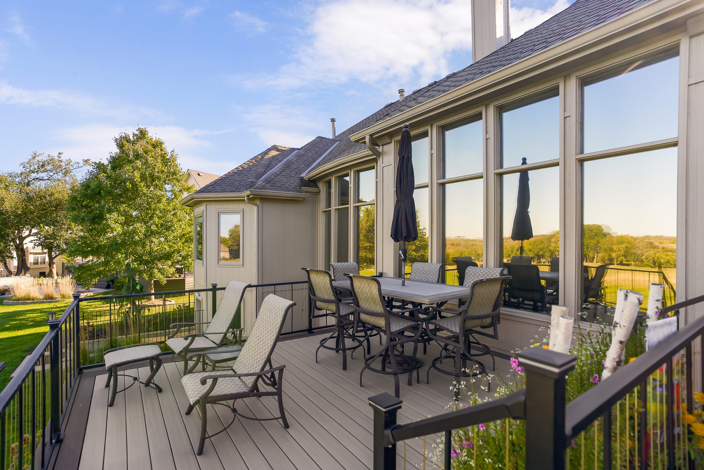 A serene deck with chairs and table, overlooking a lush garden and open fields, under a clear blue sky.