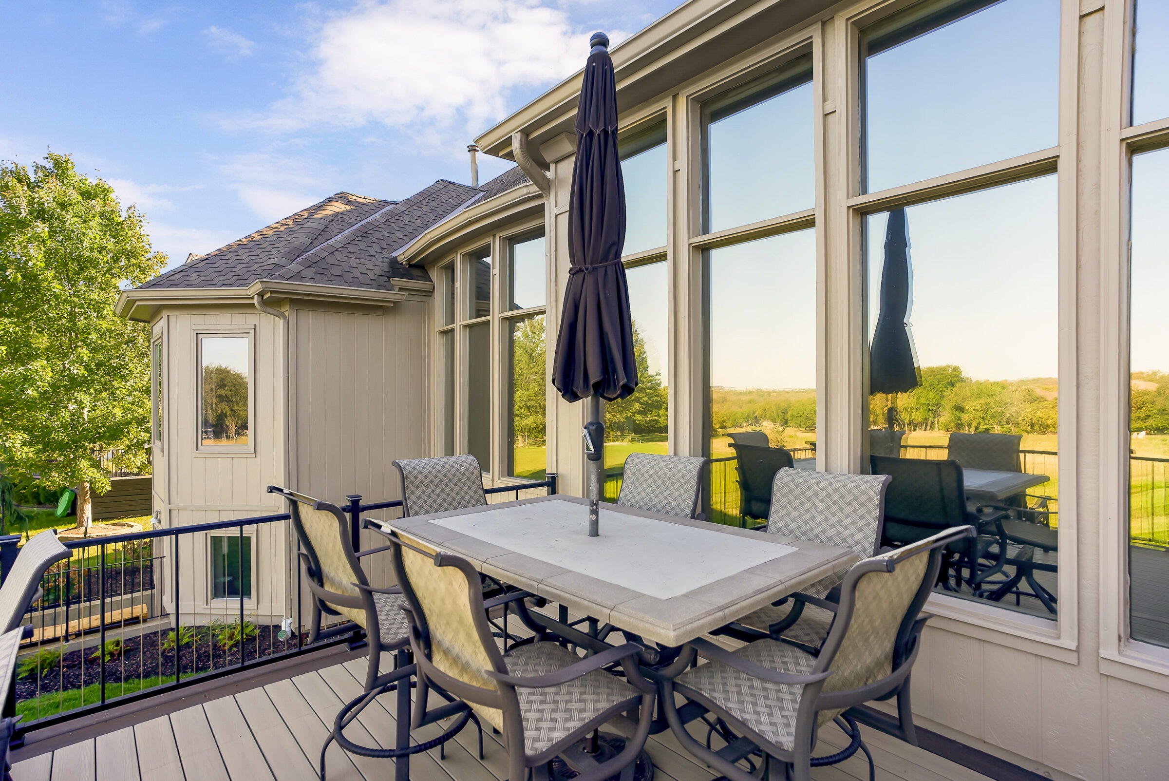 A spacious patio with outdoor seating, large windows reflecting a scenic landscape, and a closed umbrella under a clear blue sky.