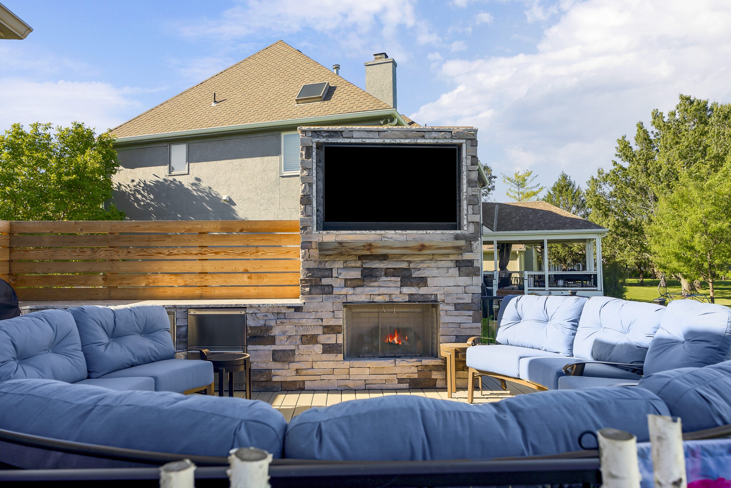 Cozy patio with blue cushioned sofas, a stone fireplace, and flat-screen TV. Wooden fence and trees create a serene backyard ambiance.